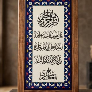 May include: A framed artwork featuring Arabic calligraphy in black ink on a cream-colored background. The frame is made of dark wood. The artwork includes decorative blue, white, red, and yellow patterned borders.