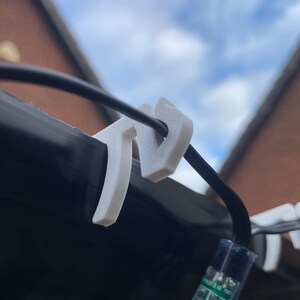 May include: White plastic clips for hanging fairy lights, attached to a black wire. The clips are shaped like the letter "V" and are holding a string of lights against a black surface.