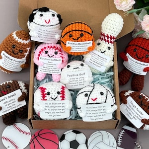 May include: A collection of handmade crochet sports balls, including footballs, basketballs, and baseballs, each with a positive message attached. The set includes a variety of sports-themed keychains. The items are displayed in a cardboard box.