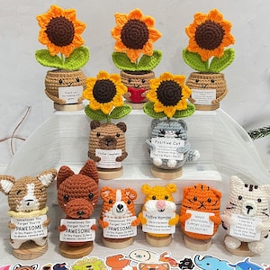 Könnte beinhalten: Eine Sammlung handgefertigter Häkelartikel, darunter Sonnenblumen, Bären, Katzen, Hunde und Hamster. Jeder Artikel steht auf einem Holzsockel und hält ein kleines Schild mit positiven Botschaften. Das Bild enthält auch einen Satz Tieraufkleber.