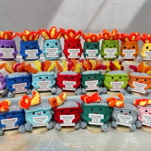 May include: A collection of colorful, crocheted dumpster figures with flames on top. Each figure has a smiling face and holds a sign that reads "Emotional Support Dumpster Fire. It's fine. Everything is fine."