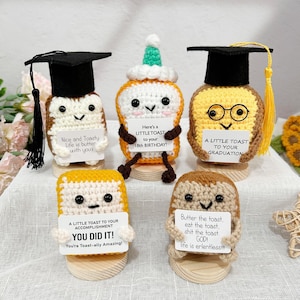 Puede incluir: Una colección de figuras de tostadas de ganchillo hechas a mano, cada una con características únicas. Algunas llevan gorros de graduación, otras sostienen carteles con mensajes como "Nice and Toasty". Las figuras de tostadas están sobre bases de madera.