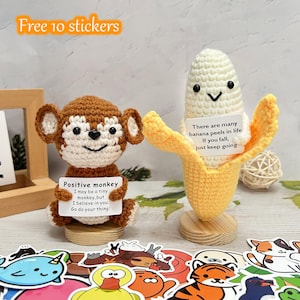 May include: Crocheted monkey and banana figurines with inspirational messages. The monkey is brown and white, holding a sign that reads "Positive monkey." The banana is yellow and white, with a sign that says "There are many banana peels in life." Includes stickers.