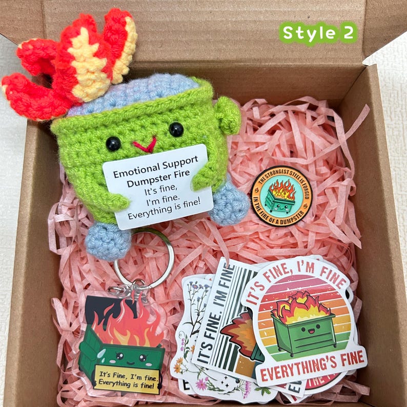 Peut inclure: Coffret cadeau avec un personnage de poubelle crochet&eacute; vert avec une flamme rouge, tenant une pancarte avec l'inscription "Emotional Support Dumpster Fire. It's fine, I'm fine, Everything is fine !" Comprend des autocollants, un porte-cl&eacute;s et une &eacute;pingle.