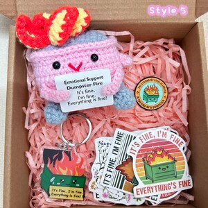 Peut inclure: Coffret cadeau avec une peluche rose en forme de feu de poubelle au crochet avec un n&oelig;ud rouge, un porte-cl&eacute;s, des autocollants et une &eacute;pingle. La peluche tient une pancarte avec l'inscription "Emotional Support Dumpster Fire".