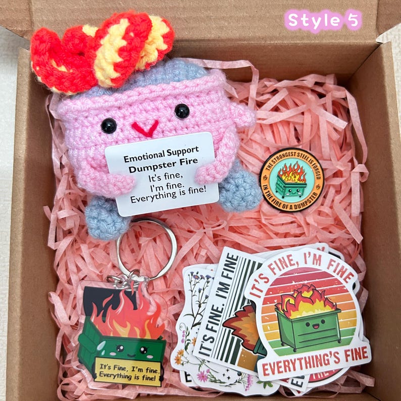 Peut inclure: Coffret cadeau avec une peluche rose en forme de feu de poubelle au crochet avec un n&oelig;ud rouge, un porte-cl&eacute;s, des autocollants et une &eacute;pingle. La peluche tient une pancarte avec l'inscription "Emotional Support Dumpster Fire".