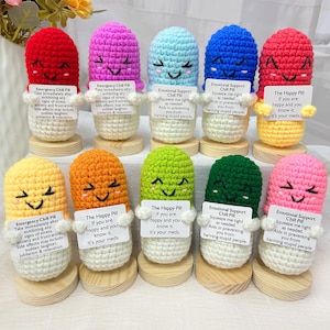 Puede incluir: Diez peluches de punto de ganchillo con forma de píldora con caras sonrientes, cada una sosteniendo un cartel con un mensaje diferente. Los mensajes incluyen "Emergency Chill Pill", "Emotional Support Chill Pill" y "The Happy Pill".