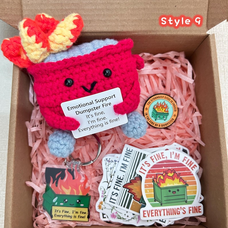 Peut inclure: Coffret cadeau avec une poubelle rouge crochet&eacute;e souriante, des autocollants, un porte-cl&eacute;s et une &eacute;pingle. La poubelle porte une pancarte avec l'inscription "Emotional Support Dumpster Fire" et la phrase "It's fine, I'm fine, everything's fine!"