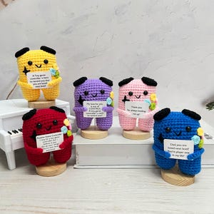 Peut inclure: Plusieurs figurines faites à la main en crochet, de couleurs jaune, rouge, violet, rose et bleu. Chaque figurine tient une petite pancarte avec du texte et un bouquet de fleurs colorées. Les figurines sont sur des socles en bois.