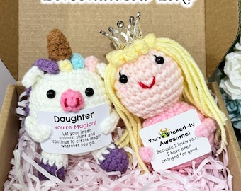 May include: Two crocheted plush toys in a box. One is a white unicorn with a brown horn and purple hooves holding a card that says Daughter You're Magical. The other is a pink doll with yellow yarn hair and a silver crown holding a card that says You're Wicked-ly Awesome!
