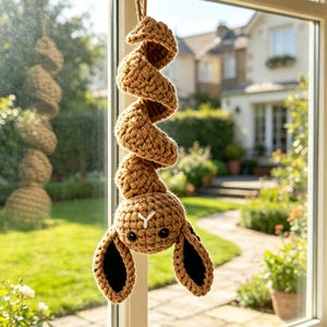 May include: A tan crochet hanging ornament featuring a spiral-shaped body and a head with two long ears. The ears have black inner sections, and the head features two small black eyes and a white stitched Y-shaped nose detail.