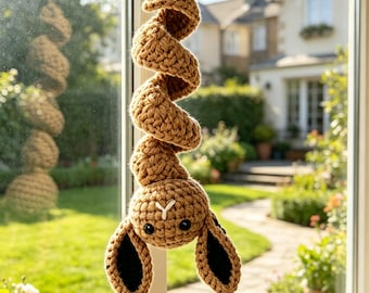May include: A tan crochet hanging ornament featuring a spiral-shaped body and a head with two long ears. The ears have black inner sections, and the head features two small black eyes and a white stitched Y-shaped nose detail.