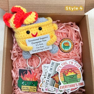 Peut inclure: Une bo&icirc;te cadeau contenant un personnage de poubelle crochet&eacute; jaune avec un n&oelig;ud rouge, tenant une pancarte qui dit "Emotional Support Dumpster Fire". Comprend des autocollants, un porte-cl&eacute;s et une &eacute;pingle sur le m&ecirc;me th&egrave;me. Le texte "It's fine, I'm fine, Everything is fine!" est visible.