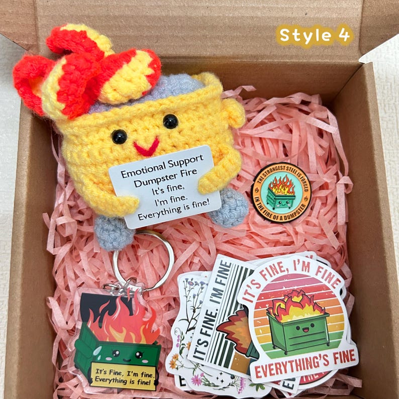 Peut inclure: Une bo&icirc;te cadeau contenant un personnage de poubelle crochet&eacute; jaune avec un n&oelig;ud rouge, tenant une pancarte qui dit "Emotional Support Dumpster Fire". Comprend des autocollants, un porte-cl&eacute;s et une &eacute;pingle sur le m&ecirc;me th&egrave;me. Le texte "It's fine, I'm fine, Everything is fine!" est visible.