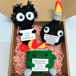 May include: A handmade crochet gift set featuring three whimsical characters: a black creature, a bomb, and a green dumpster fire. Each character holds a sign with the phrase "It's fine, I'm fine, Everything is fine!" The bomb reads "Feeling stressed? Squeeze tightly, aim and release. Satisfaction guaranteed!"