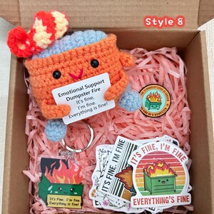 Peut inclure: Une bo&icirc;te cadeau avec un jouet en crochet orange en forme de benne &agrave; ordures avec un chapeau rouge et blanc, tenant une pancarte avec l'inscription "Emotional Support Dumpster Fire". Comprend des autocollants, un porte-cl&eacute;s et une &eacute;pingle.