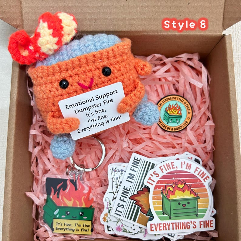 Peut inclure: Une bo&icirc;te cadeau avec un jouet en crochet orange en forme de benne &agrave; ordures avec un chapeau rouge et blanc, tenant une pancarte avec l'inscription "Emotional Support Dumpster Fire". Comprend des autocollants, un porte-cl&eacute;s et une &eacute;pingle.