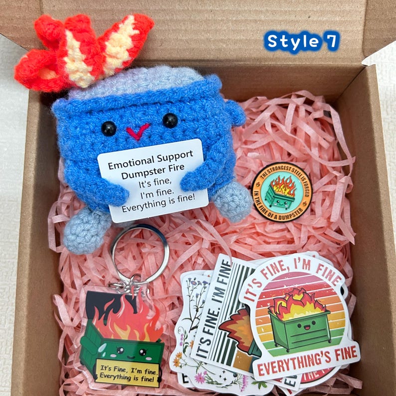 Peut inclure: Un jouet de poubelle bleu au crochet avec un accent en forme de flamme rouge et jaune, un porte-cl&eacute;s et des autocollants, tous avec la phrase "It's fine, I'm fine, Everything is fine!". Les articles sont dans une bo&icirc;te en carton avec du papier rose.