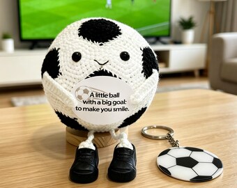 May include: A crocheted soccer ball plush with black and white pentagon patterns, black eyes, and a small smile. It holds a circular white sign with the text: A little ball with a big goal: to make you smile. It has white legs and black shoes.