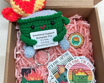May include: A crochet dumpster toy with a red and yellow flame design, a keychain, and stickers. The dumpster has a sign that reads "Emotional Support Dumpster Fire. It's fine, I'm fine. Everything is fine!" The items are in a cardboard box.