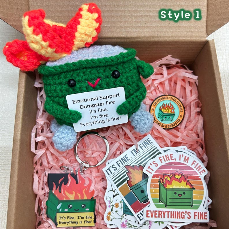 Peut inclure: Un jouet de poubelle au crochet avec un motif de flamme rouge et jaune, un porte-cl&eacute;s et des autocollants. La poubelle porte une pancarte indiquant "Emotional Support Dumpster Fire. It's fine, I'm fine. Everything is fine !" Les articles sont dans une bo&icirc;te en carton.