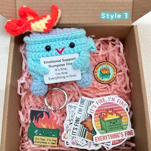 Peut inclure: Une bo&icirc;te cadeau contenant une poubelle bleue crochet&eacute;e avec une flamme rouge et jaune, un porte-cl&eacute;s, des autocollants et une &eacute;pingle. La poubelle porte une pancarte avec le texte "Emotional Support Dumpster Fire. It's fine, I'm fine. Everything is fine!"