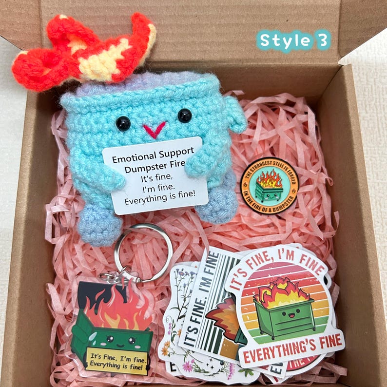 Peut inclure: Une bo&icirc;te cadeau contenant une poubelle bleue crochet&eacute;e avec une flamme rouge et jaune, un porte-cl&eacute;s, des autocollants et une &eacute;pingle. La poubelle porte une pancarte avec le texte "Emotional Support Dumpster Fire. It's fine, I'm fine. Everything is fine!"