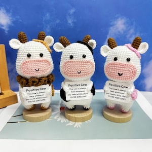 Puede incluir: Tres figuritas de vaca de crochet blanco con detalles marrones, negros y rosas. Cada vaca sostiene un cartel que dice "Positive Cow Tiny cow is always here whenever you need a little cheer. YOU'RE AWESOME!"