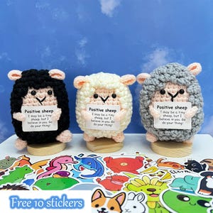 May include: Three crocheted sheep toys in black, cream, and grey, each holding a sign with the text "Positive sheep." The sheep are on wooden bases, with a collection of colourful stickers in the foreground. The text on the sign reads: "I may be a tiny sheep, but I believe in you. Go do your thing!"