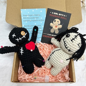 May include: A voodoo doll set presented in a cardboard box. The set features two crocheted dolls, one black with a red heart and one off-white. The box also contains pins and cards with text about the dolls.