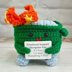 Op de afbeelding: Een groene gehaakte vuilnisbakbrand met een blauw gehaakt deksel en oranje gehaakte vlammen. De vuilnisbakbrand houdt een bord vast met de tekst "Emotional Support Dumpster Fire It's fine, I'm fine. Everything is fine!"