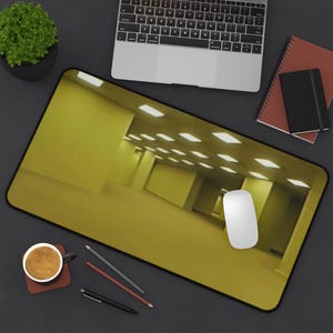 May include: A rectangular desk mat featuring a printed image of a yellow, dimly lit, empty office hallway with fluorescent ceiling lights. The mat is positioned on a dark surface alongside a laptop, a coffee mug, a computer mouse, and pens.