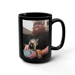 Peut inclure: Un mug en céramique noire présentant une photographie imprimée d'une personne aux cheveux bouclés et à la barbe, portant un bonnet, un t-shirt noir à motif et une pile de bracelets en perles colorées, tenant un bol bleu clair.