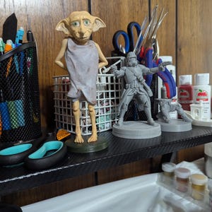 May include: A shelf displays several figurines, including a Dobby figure and a Star Wars Stormtrooper. The Dobby figure is light-skinned with large ears and is wearing a gray cloth. The Stormtrooper is gray and in a battle pose. Other items include scissors and paint.