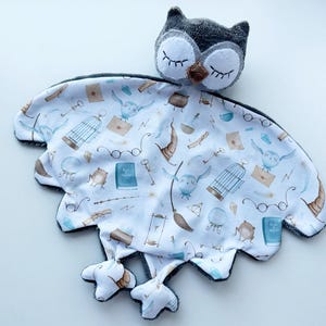 May include: A plush owl-shaped baby security blanket. The owl head is gray with white accents and closed eyes. The blanket is white with a pattern of owls, books, and other magical elements in blue and brown. The blanket has a scalloped edge.