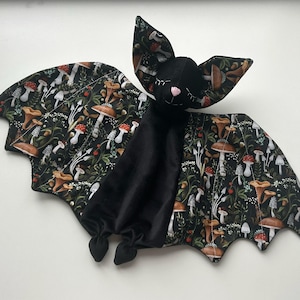 May include: A plush bat toy with black velvet body and mushroom-patterned wings. The wings feature a dark background with illustrations of various mushrooms and foliage. The bat has a pink nose and closed eyes.