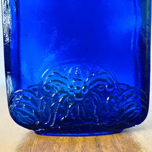 May include: A cobalt blue glass bottle with a decorative floral design on the bottom. The bottle has a rectangular shape and is sitting on a wooden surface.