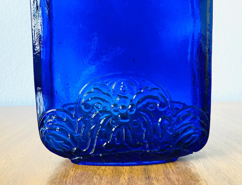 May include: A cobalt blue glass bottle with a decorative floral design on the bottom. The bottle has a rectangular shape and is sitting on a wooden surface.