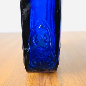 May include: A cobalt blue glass bottle with a decorative embossed design. The bottle has a rectangular shape with rounded corners and a dark blue, almost black, border. The design is visible on the front of the bottle.