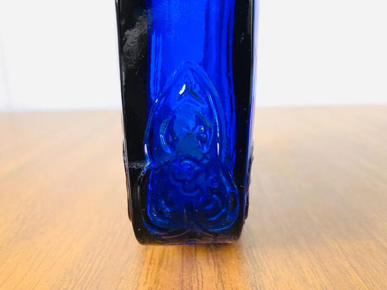 May include: A cobalt blue glass bottle with a decorative embossed design. The bottle has a rectangular shape with rounded corners and a dark blue, almost black, border. The design is visible on the front of the bottle.