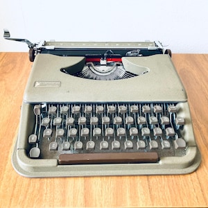 Könnte beinhalten: Eine olivgrüne Vintage-Schreibmaschine mit voller Tastatur und Chromakzenten steht auf einem Holztisch. Die Tasten der Schreibmaschine sind grau, und der Wagenrücklaufhebel ist sichtbar. Die Maschine ist ein klassisches Beispiel für Büroausstattung aus der Mitte des Jahrhunderts.