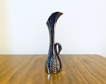 May include: A metallic silver swan-shaped vase with a curved neck and a swan's head and neck forming the handle. The vase is on a wooden surface against a white wall. The vase is approximately 10 inches tall.