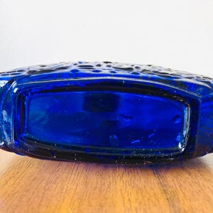 May include: A cobalt blue glass bottle with a rectangular shape and textured surface. The bottle is lying on a wooden surface. The bottle's design features a textured pattern on the top.