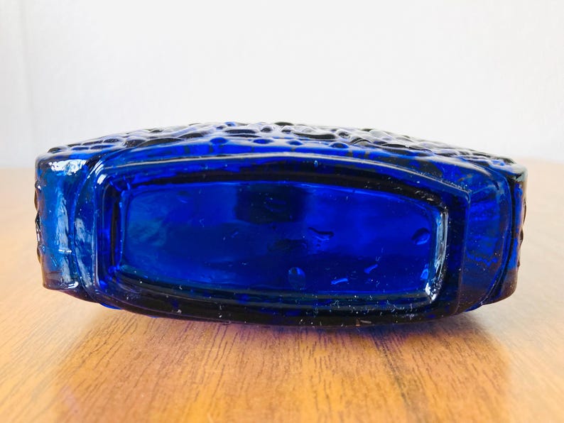 May include: A cobalt blue glass bottle with a rectangular shape and textured surface. The bottle is lying on a wooden surface. The bottle's design features a textured pattern on the top.