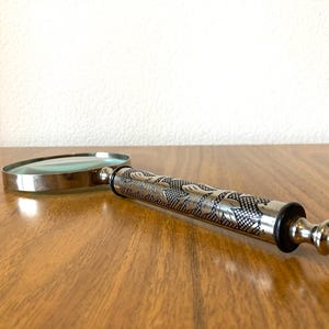 May include: A silver-toned magnifying glass with an ornate, engraved handle. The handle features a detailed pattern, and the glass lens is clear. The magnifying glass is resting on a wooden surface.