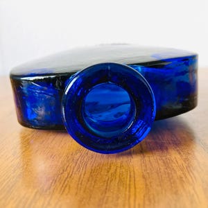 May include: A cobalt blue glass bottle with a rounded rectangular shape. The bottle has a wide, circular opening and a flat top. The glass has a glossy finish, and the bottle is sitting on a wooden surface.