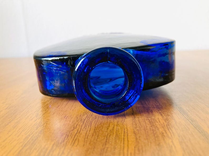 May include: A cobalt blue glass bottle with a rounded rectangular shape. The bottle has a wide, circular opening and a flat top. The glass has a glossy finish, and the bottle is sitting on a wooden surface.