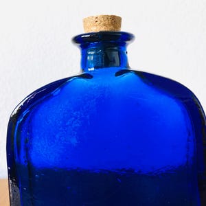 May include: A cobalt blue glass bottle with a cork stopper. The bottle has a square shape with rounded edges and a textured surface. The cork is a light brown color.