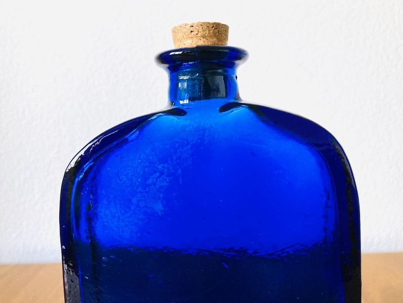 May include: A cobalt blue glass bottle with a cork stopper. The bottle has a square shape with rounded edges and a textured surface. The cork is a light brown color.