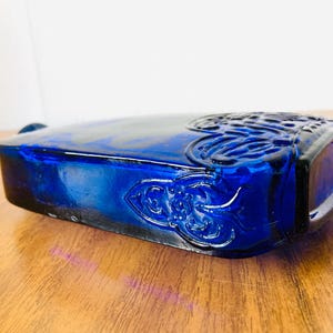 May include: A rectangular cobalt blue glass bottle with ornate embossed floral and scroll designs. The bottle has a rounded top and is resting on a wooden surface.